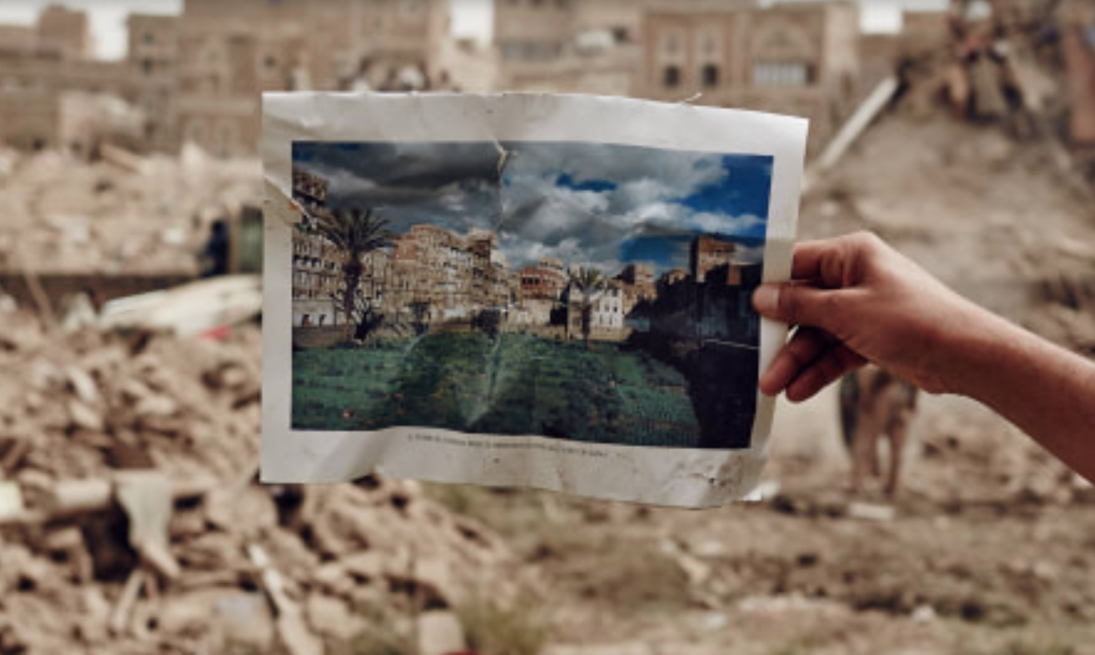 A hand holds a photograph of a home before a pile of rubble.