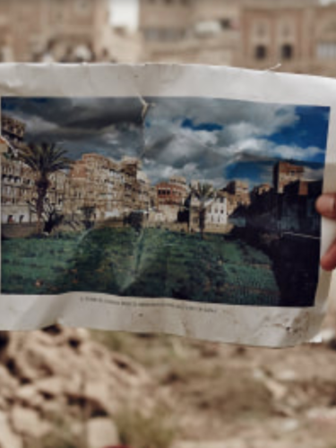 A hand holds a photograph of a home before a pile of rubble.